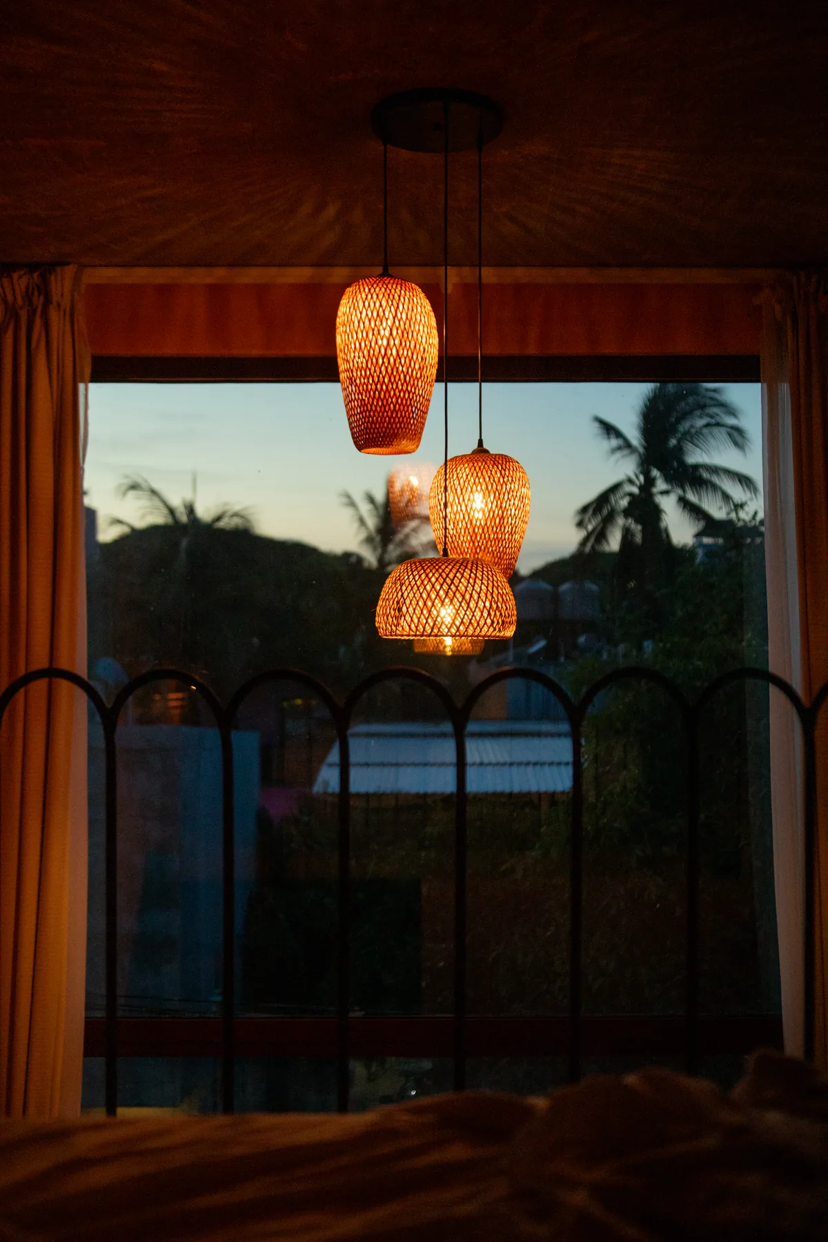 Woven pendant lamps glowing over a bed at dusk, with palm silhouettes through the window at Amplitude Coliving, La Punta